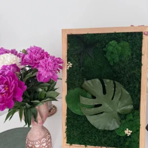 Mechový obraz s listem Monstera v dřevěném rámu, umístěný vedle vázy s růžovými pivoňkami.